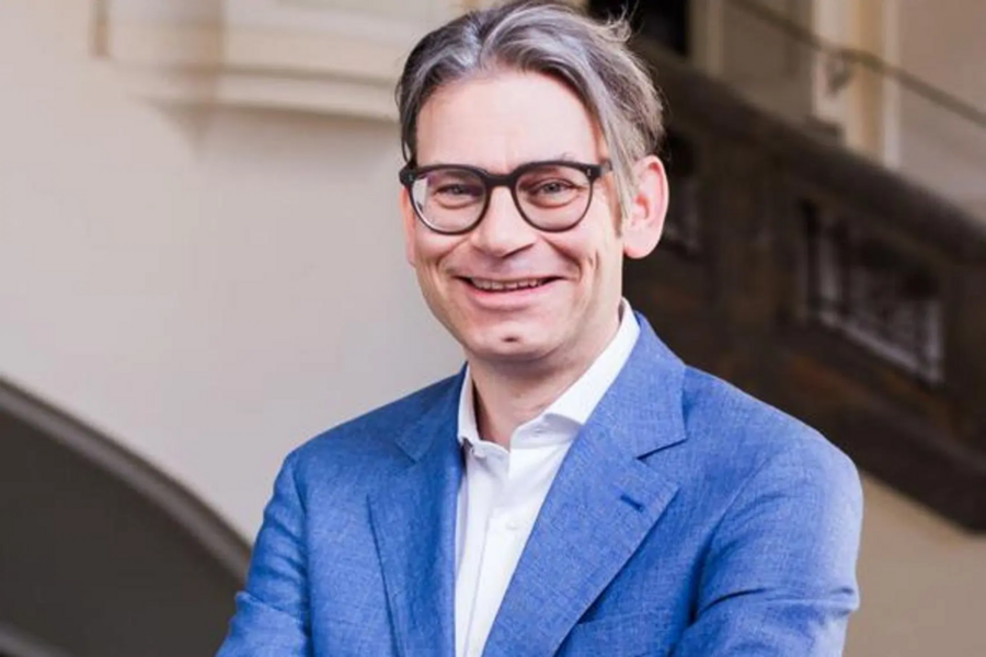 A man wearing glasses and a blue suit, smiles while standing indoors in front of a staircase.