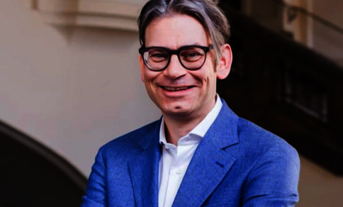 Ein Mann mit Brille und blauem Anzug lächelt, während er drinnen vor einer Treppe steht.