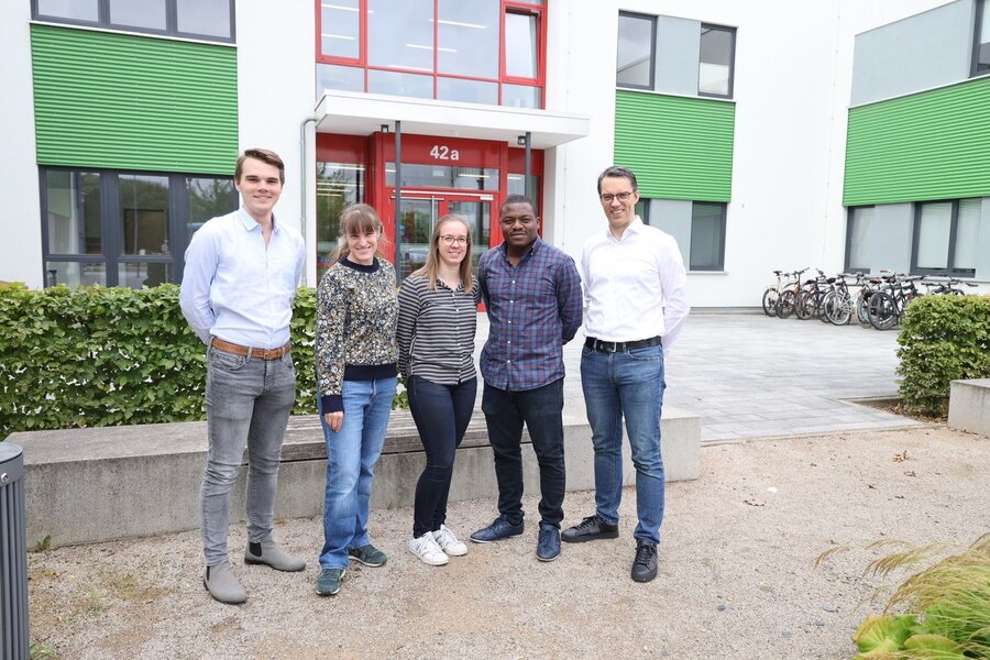 Five people stand in front of a building and smile at the camera. The building has white walls and green shutters, the surroundings are well-tended with plants and a paved path. 