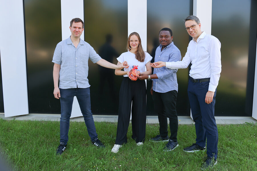 Four people stand on grass in front of a modern building, holding a model of a human heart together and facing the camera.