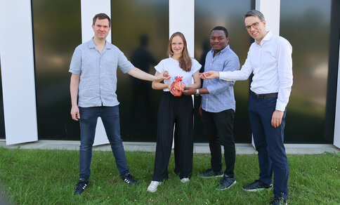 Four people stand on grass in front of a modern building, holding a model of a human heart together and facing the camera.
