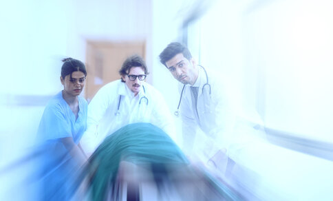 Bewegungsunscharfe Aufnahme eines medizinischen Teams, das einen Patienten auf einer Trage durch einen hellen Krankenhausflur schiebt. Das Bild vermittelt Dringlichkeit und die Situation einer medizinischen Notfallversorgung.