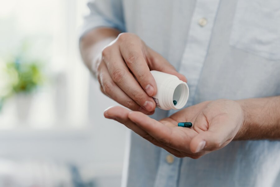 A close-up of a person wearing a light grey shirt, holding a small white medicine bottle and pouring tablets into their open hand.
