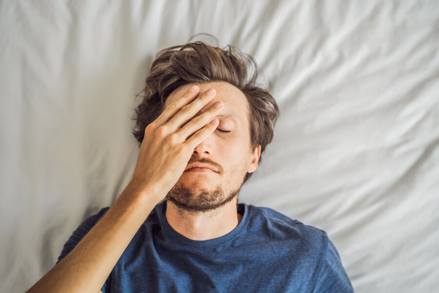 A man lying on his back in bed with his eyes closed, resting his hand over his forehead in exhaustion, wearing a blue T-shirt.
