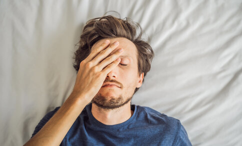 A man lying on his back in bed with his eyes closed, resting his hand over his forehead in exhaustion, wearing a blue T-shirt.