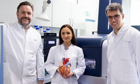 In a laboratory, three researchers in lab coats are looking at the camera. Dr Aiste Liutkute stands between Dr Christof Lenz and Prof. Niels Voigt, holding a model heart up to the camera.