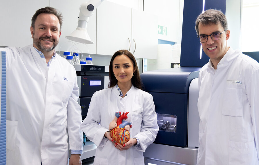 In a laboratory, three researchers in lab coats are looking at the camera. Dr Aiste Liutkute stands between Dr Christof Lenz and Prof. Niels Voigt, holding a model heart up to the camera.