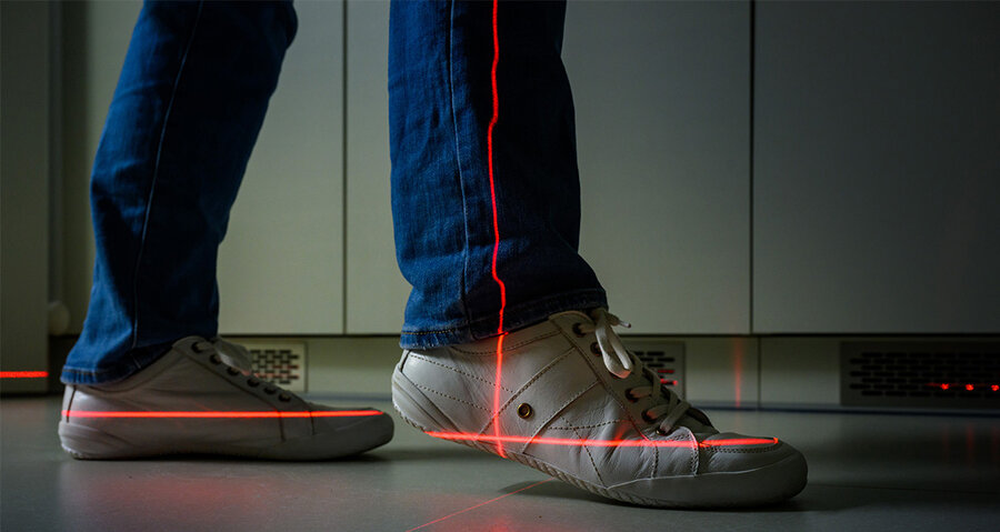 Person wears white trainers and blue jeans, red laser beam runs along the right shoe on the floor