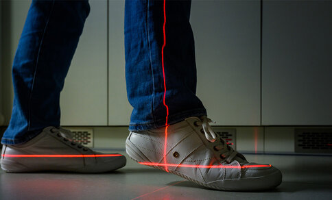 Person trägt weiße Sneaker und blaue Jeans, roter Laserstrahl verläuft entlang des rechten Schuhs auf dem Boden