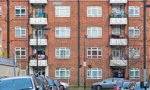 Red brick building with flats, cars parked on the street in front of it. 