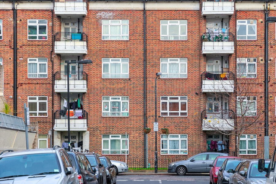 Red brick building with flats, cars parked on the street in front of it. 