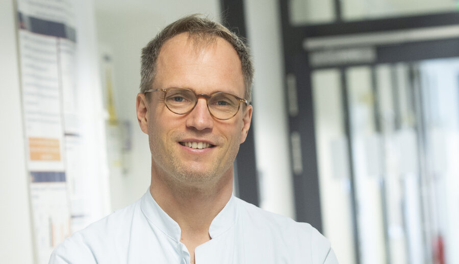  A man with short, slightly wavy, light brown hair and glasses with thin, brown frames stands in a brightly lit corridor. He is wearing a white medical top and smiles friendly into the camera. Scientific posters and glass doors can be seen in the background, suggesting a research facility or hospital.