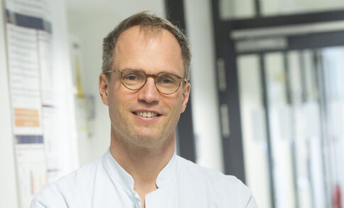  A man with short, slightly wavy, light brown hair and glasses with thin, brown frames stands in a brightly lit corridor. He is wearing a white medical top and smiles friendly into the camera. Scientific posters and glass doors can be seen in the background, suggesting a research facility or hospital.