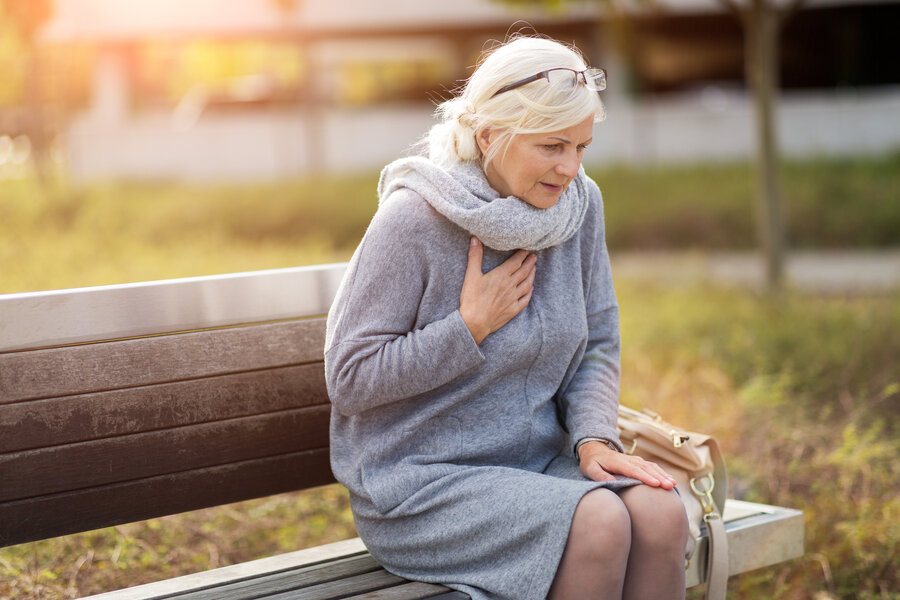 An elderly woman is sitting on a park bench and has chest pains.