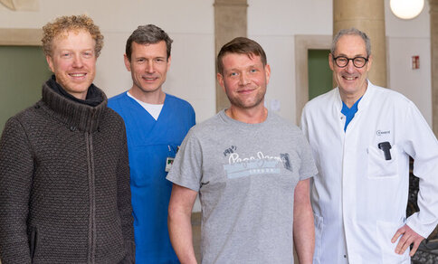 Four men stand next to each other, two in medical clothing, two in casual wear, smiling at the camera