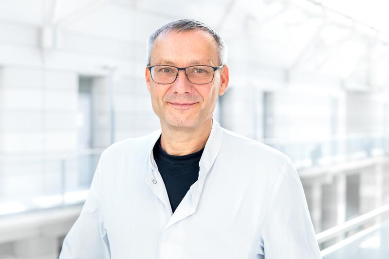 A man with glasses ( he is Prof. Dr Frank Edelmann) in a white doctor's coat stands smiling in a brightly lit, modern building.