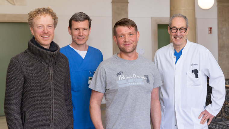 Four men stand next to each other, two in medical clothing, two in casual wear, smiling at the camera
