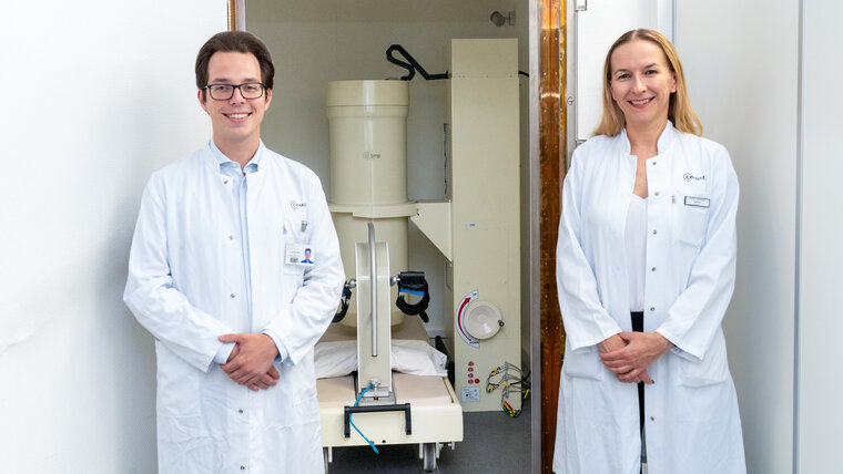 Two researchers in white lab coats stand smiling in the doorway of an examination room. A magnetocardiograph can be seen in the background.