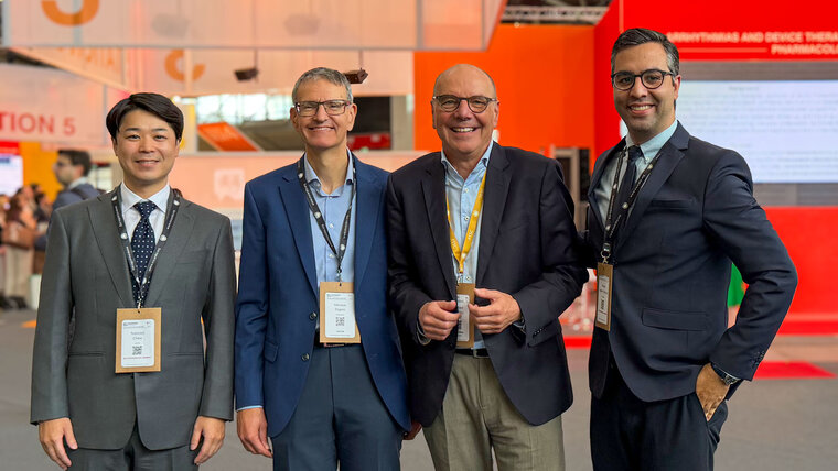 Four men in suits are standing at a trade fair. They are smiling and wearing name badges. In the background, blurred stands and displays can be seen, conveying the atmosphere of a business event.