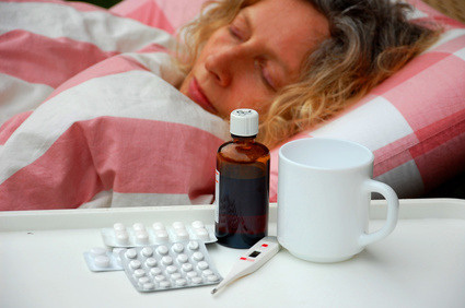 Erkrankte Frau liegt im Bett, auf dem Nachttisch stehen Medikamente, ein Thermometer und eine Tasse.