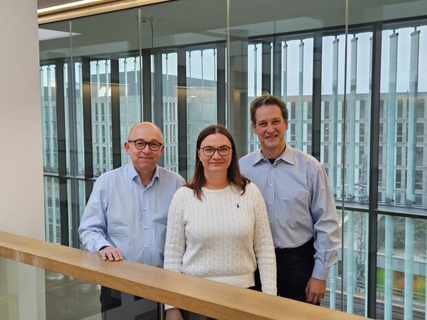 Drei Personen in einem modernen Gebäude mit Glasfassade: links ein Mann(Prof. Dr. med. Christoph Knosalla), eine Frau(Dr. Mariya Kucherenko ) in der Mitte und ein Mann rechts( Prof. Dr. med. Wolfgang Kübler), alle in hellblauer Kleidung und weißem Pullover.